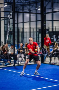 Riga, Letonya - 8 Aralık 2024 - Kırmızı tişörtlü ve siyah şortlu bir erkek padel oyuncu, kapalı bir mavi padel kortunda seyircilerin gözü önünde topa vurmaya hazırlanıyor.