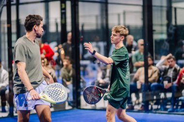 Riga, Letonya - 8 Aralık 2024 - Maç sırasında sahada iki Padel oyuncusu, biri diğerine doğru elini uzatıyor. Seyirciler camın arkasında görünür. Padel.