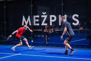 Riga, Letonya - 8 Aralık 2024 - 2 padel oyuncu kapalı sahada iş başında. Kırmızılı bir oyuncu topa hamle yaparken siyahlı olan cevap vermeye hazırlanıyor. Padel.