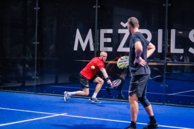 Riga, Letonya - 8 Aralık 2024 - kapalı sahada iki erkek padel oyuncusu. Biri kırmızı dalışlar yaparken, diğeri dikkatle izliyor. Padel 'de eylem dolu bir an.