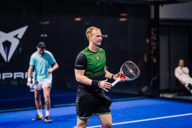 Riga, Letonya - 8 Aralık 2024 - kapalı sahada iki erkek padel oyuncusu. Yeşil gömlekli biri harekete hazırlanırken, diğeri arkada deniz mavisi renginde bekliyor. Padel sporu.