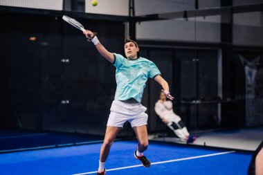 Riga, Letonya - 8 Aralık 2024 - Deniz mavisi tişörtlü bir erkek, kapalı sahada oynanan bir maçta topa forehand salıncağıyla vurmak için atlar. Padel spor hareketi.