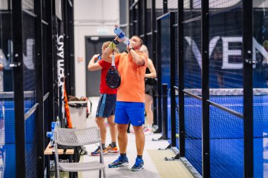 Riga, Letonya - 8 Aralık 2024 - Turuncu tişörtlü bir padel oyuncusu mola sırasında elinde raketle suyla su içer. Ekipmanlı kapalı padel kort ayarları.