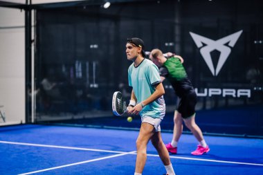 Riga, Letonya - 8 Aralık 2024 - Biri topa vurmaya hazırlanırken diğeri arka planda oynayan iki padel oyuncusu iç sahada mavi sahada hareket halinde. Padel eşleşmesi