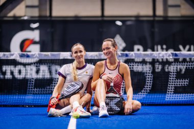 Riga, Letonya - 8 Aralık 2024 - İki bayan Padel oyuncusu arka planda ağ ve spor markalarıyla mavi kortta oturmuş gülümsüyor. Padel eşleştirme sahnesi