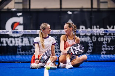 Riga, Letonya - 8 Aralık 2024 - İki gülümseyen bayan Padel oyuncusu, arka planda ağ ve spor markalarıyla mavi bir kortta oturuyor. Padel eşleştirme sahnesi.