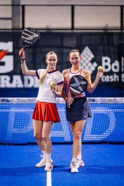 Riga, Letonya - 8 Aralık 2024 - Spor kıyafetleri içinde iki bayan padel oyuncusu, bir maçtan sonra güvenle gülümseyen padel raket ve toplarla mavi bir kortta duruyorlar. Padel.