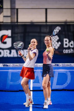 Riga, Letonya - 8 Aralık 2024 - Spor kıyafetleri içinde iki bayan padel oyuncusu, bir maçtan sonra güvenle gülümseyen padel raket ve toplarla mavi bir kortta duruyorlar. Padel.