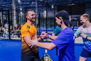 Riga, Letonya - 8 Aralık 2024 - Dört Padel oyuncusu mavi sahada el sıkışıyor ve gülümsüyor, maçtan sonra, yoldaşlık ve sportmenlik ile kapalı bir arenada kutlama yapıyorlar..