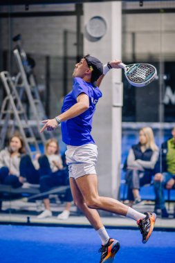 Riga, Letonya - 8 Aralık 2024 - Mavi tişörtlü bir erkek padel oyuncusu raketiyle topa vurmak için havaya sıçrıyor, kapalı bir mavi kortta odak ve atletizm gösteriyor..