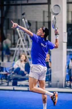 Riga, Letonya - 8 Aralık 2024 - Mavi tişörtlü bir erkek padel oyuncusu maç sırasında topa raketiyle vurmak için atlıyor, mavi bir korta odaklanma ve atletizm gösteriyor..