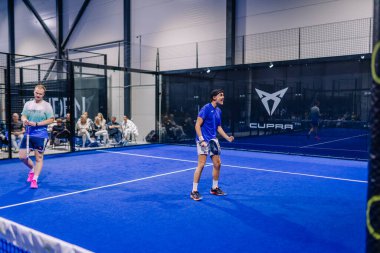 Riga, Letonya - 8 Aralık 2024 - Mavi kortta iki padel oyuncusu, biri coşkuyla bir anı kutlarken diğeri kapalı bir arenada seyirciler tarafından çevrili.