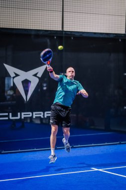 Riga, Letonya - 8 Aralık 2024 - Bir erkek padel oyuncusu elinde raketle topa vurmak için havadan atlıyor, mavi bir korta odaklanmış bir ifadeyle, Cupra logosunun yer aldığı.