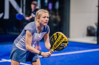 Riga, Letonya - 8 Aralık 2024 - Odaklanmış bir bayan padel oyuncusu, mavi sahada ağır bir maç sırasında sarı bir Babolat raketi tutarak topu iade etmeye hazırlanıyor..