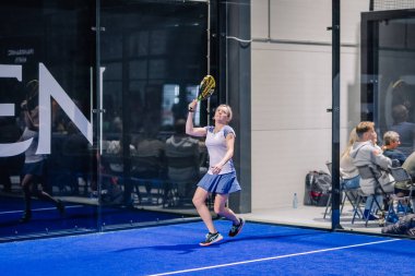 Riga, Letonya - 8 Aralık 2024 - Bir bayan padel oyuncusu, kapalı bir spor tesisinin cam duvarından izleyen seyircilerle mavi kortta topa vurmaya hazırlanıyor..
