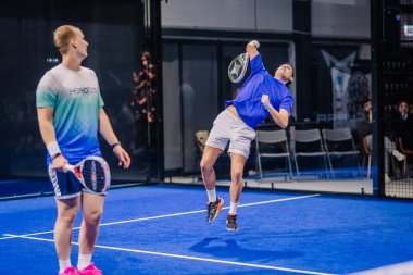 Riga, Letonya - 8 Aralık 2024 - Mavi sahada iki padel oyuncusu, yoğun bir maç sırasında güçlü bir atış için atlayan bir oyuncu.