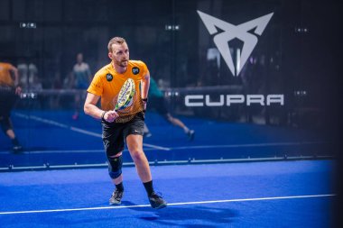Riga, Letonya - 8 Aralık 2024 - Mavi kortta raket tutan ve maç sırasında cam duvarında bir Cupra logosu olan bir padel oyuncusu.
