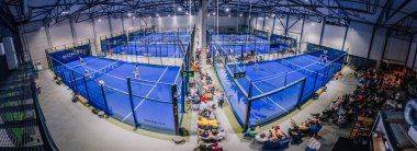 Riga, Letonya - 8 Aralık 2024 - Birden fazla maç yapılan kapalı padel mahkemeler, modern bir spor tesisinin saha kenarından maç izleyen seyirciler.