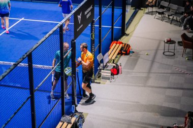 Riga, Letonya - 8 Aralık 2024 - İki Padel oyuncusu internetin yakınında etkileşim halindeler. Banklar, spor malzemeleri ve bir dizüstü bilgisayar mahkeme dışında görülebiliyor. Padel eşleşme ayarları.