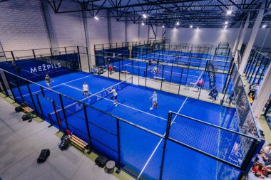 Riga, Letonya - 8 Aralık 2024 - Yüksek çitlerle çevrili mavi kortlarda aktif olarak maç yapan oyuncuların bulunduğu kapalı bir padel saha tesisinin havadan görüntüsü.