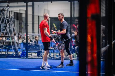 Riga, Letonya - 8 Aralık 2024 - İki Padel oyuncusu kapalı bir sahada tartışıyorlar. Biri dinlerken diğeri bir padel raketi tutuyor. İzleyiciler arka planda oturur.