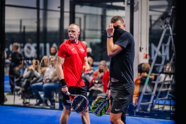 Riga, Letonya - 8 Aralık 2024 - İki erkek Padel oyuncusu kapalı sahada mola veriyor; biri raketini tutarken diğeri yüzünü siliyor. Padel eşleşmesi devam ediyor.