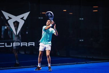 Riga, Letonya - 8 Aralık 2024 - Turkuaz tişörtlü ve beyaz şortlu bir erkek padel oyuncusu kapalı bir mavi kortta raketiyle topa vurmaya hazırlanıyor. Padel.