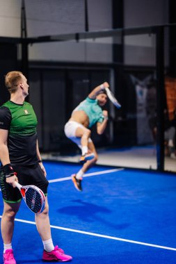 Riga, Letonya - 8 Aralık 2024 - İki padel oyuncu kapalı bir mavi kortta, biri havada yüksek bir vuruş yapmaya hazırlanırken diğeri ise bir padel raket tutuyor. Padel.