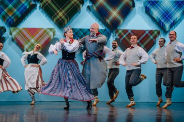 Valmiera, Letonya - 8 Aralık 2024 - Renkli geleneksel kostümlü Letonyalı halk dansçıları parlak ışıklar altında sahne alarak, senkronize hareketler yoluyla kültürel mirası sergilediler..