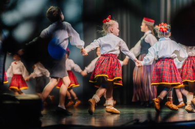 Valmiera, Letonya - 8 Aralık 2024 - Geleneksel Letonya Halk dansları kostümlü çocuklar sahne arkasında renkli eşarplar eşliğinde kollarını kaldırarak gösteri yapıyorlar..