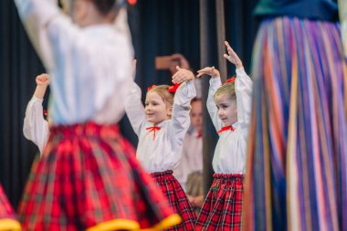 Valmiera, Letonya - 8 Aralık 2024 - Genç bir çocuk Letonya Halk danslarında kolları havada, sahnede geleneksel kırmızı ve sarı kostümlü diğer çocuklar tarafından sarılmış bir şekilde sahneye çıkıyor..