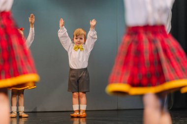 Valmiera, Letonya - 8 Aralık 2024 - Genç bir çocuk Letonya Halk danslarında kolları havada, sahnede geleneksel kırmızı ve sarı kostümlü diğer çocuklar tarafından sarılmış bir şekilde sahneye çıkıyor..