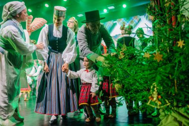 Valmiera, Latvia - December 8, 2024 - People in traditional Latvian folk costumes celebrate around a decorated Christmas tree on stage during a cultural performance. Latvian Folk dance. clipart