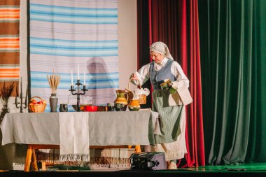 Valmiera, Letonya - 8 Aralık 2024 - Geleneksel Letonya halk kıyafetleri içindeki bir kadın kültürel bir gösteri sırasında örülmüş tekstil ve aksesuarlarla çevrili bir masada eşyalar düzenliyor..