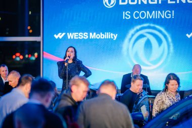 Riga, Latvia - December 2, 2024 - Singer in a sparkly dress performing on stage with a live band, vibrant lighting, and WESS Mobility branding in the background at a Dongfeng event clipart