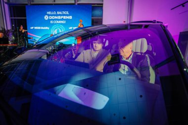 Riga, Latvia - December 2, 2024 - Two men seated inside a Dongfeng electric car with reflections on the windshield, showcased at a promotional event under vibrant lighting. clipart