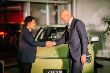 Riga, Letonya - 2 Aralık 2024 İcra Kurulu Başkanları, ortaklık ve otomotiv yeniliğini simgeleyen şirket etkinliğinde yeşil bir Dongfeng Kutusu elektrikli arabasının önünde el sıkışıyor..