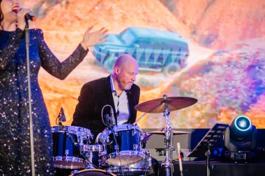 Riga, Latvia - December 2, 2024 - A drummer performs on stage with a vibrant backdrop during the Dongfeng event. The scene features a live band and colorful stage lighting. clipart