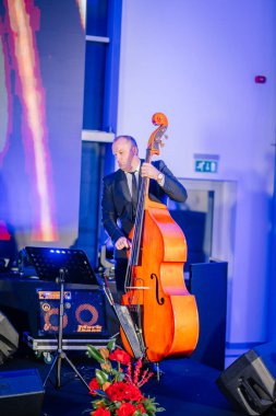 Riga, Latvia - December 2, 2024 - A male musician plays an upright bass on stage at the Dongfeng event, with vibrant lighting, floral decor, and sound equipment in the background. clipart