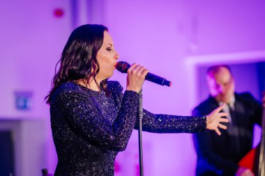 Riga, Letonya - 2 Aralık 2024 - Sahnede tutkulu bir şekilde bir mikrofonla performans sergileyen kadın şarkıcı, ışıl ışıl bir elbise giyiyor ve yanında da bir çift basçı bulunuyor. Dongfeng etkinliği.