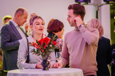 Riga, Letonya - 2 Aralık 2024 - Zarif bir dekor, çiçekler ve şarap kadehleriyle resmi bir etkinlikte şık bir kadın ve erkek sohbete girişir.