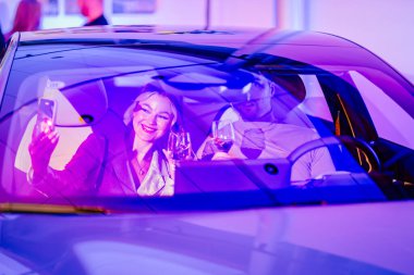 Riga, Letonya - 2 Aralık 2024 - Gülümseyen bir kadın Dongfeng arabasında bir adamla selfie çekiyor, her ikisi de şarap kadehleri tutuyor, canlı mor ışıkla aydınlatılıyor..