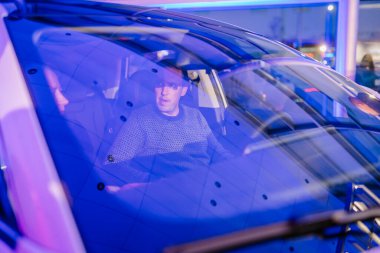 Riga, Letonya - 2 Aralık 2024 - Modern bir otomotiv etkinliğinde bir arabanın içinde oturan, ön camdan mavi yansımalı bir adam.