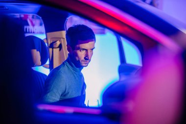 Riga, Letonya - 2 Aralık 2024 - Mavi kazaklı bir adam bir arabanın içinde oturuyor, canlı bir mavi ve kırmızı ışıkla aydınlanıyor, elinde bir bardak tutuyor..