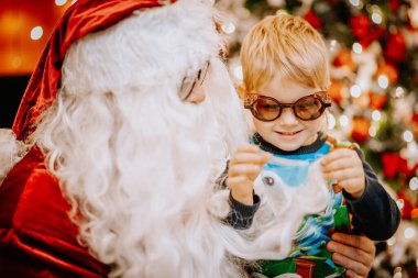 Noel Baba ve gözlüklü gülümseyen bir çocuk, Noel Baba 'nın sakalını neşeli bir şekilde inceliyor.