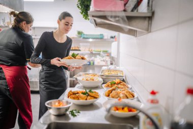 Valmiera, Letonya - 13 Aralık 2024 - Profesyonel bir mutfakta yemek düzenlerken bir tabak kızarmış tavuğu dikkatle tutan bir şef, takım çalışmasını ve hazırlıklarını vurguladı.
