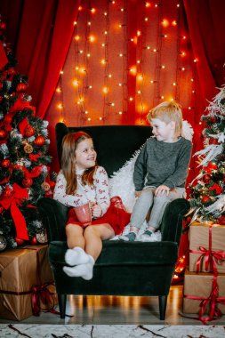 Gülümseyen bir kız ve oğlan Noel süsleriyle çevrili bir sandalyede oturuyor, parlayan ışıklar, ve festival ortamında hediyeler paketliyor..