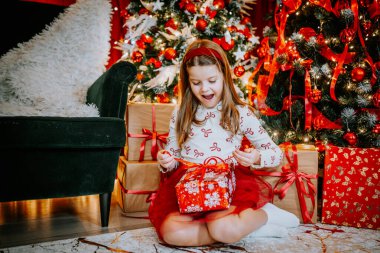 A young girl sitting near a Christmas tree, untying the ribbon on a red gift box, surrounded by festive decorations and wrapped presents. clipart