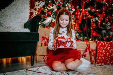 Noel ağacının yanında oturan, elinde Noel süslemeleriyle sarılı bir hediyeyle gülümseyen, neşeli bir genç kız..