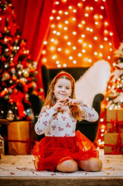 Parlayan ışıklar, ağaçlar ve paketlenmiş hediyelerle, neşeli bir Noel ortamında oturmuş, elleriyle kalp şekli oluşturan neşeli bir kız..